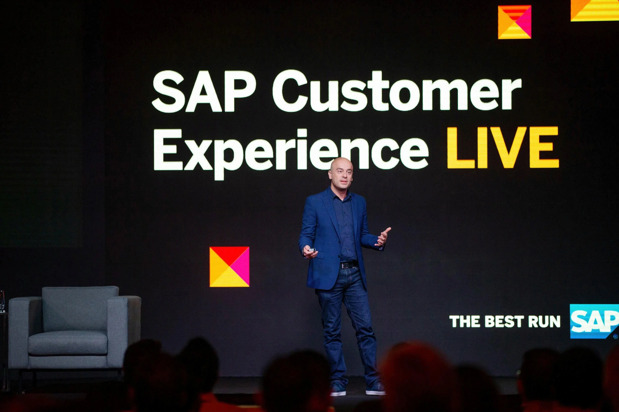 A man in a blue blazer and jeans speaking on stage at a conference. Behind him is a large screen with the text 'SAP Customer Experience LIVE' and the SAP logo, with a small colorful logo in the top right corner and bottom right corner.