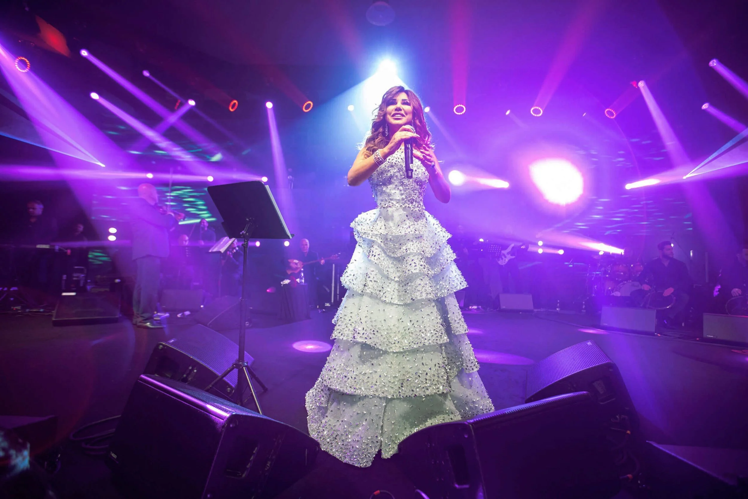 A woman in a white, tiered, beaded gown performing on stage with purple and pink lights, holding a microphone, with a band in the background.