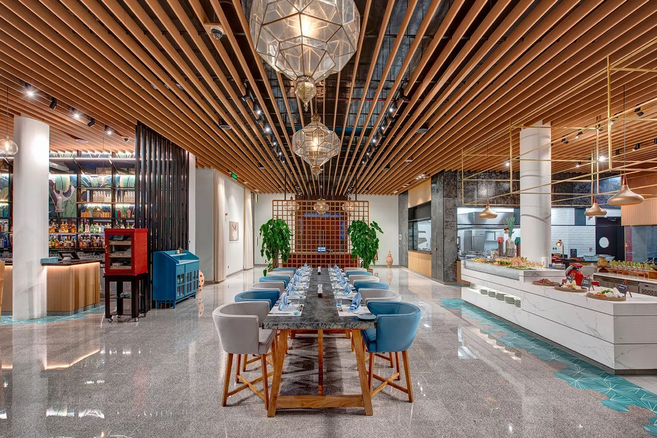 Interior of a modern, upscale restaurant with a large dining table set with place settings, surrounded by chairs, and decorative greenery.