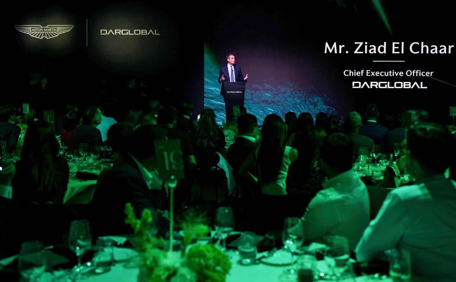 A man in a suit giving a presentation on stage at a conference, with a large screen behind him displaying the name Mr. Ziad El Chaar, Chief Executive Officer of DARGLOBAL, and the DARGLOBAL logo. The audience is seated at tables with drinks and table
