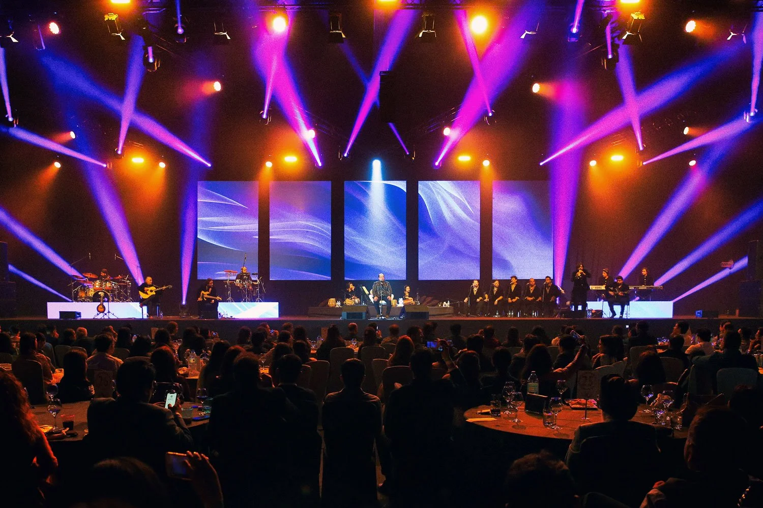 A concert stage with musicians performing under colorful purple, orange, and blue spotlights, with a large audience seated at tables watching.