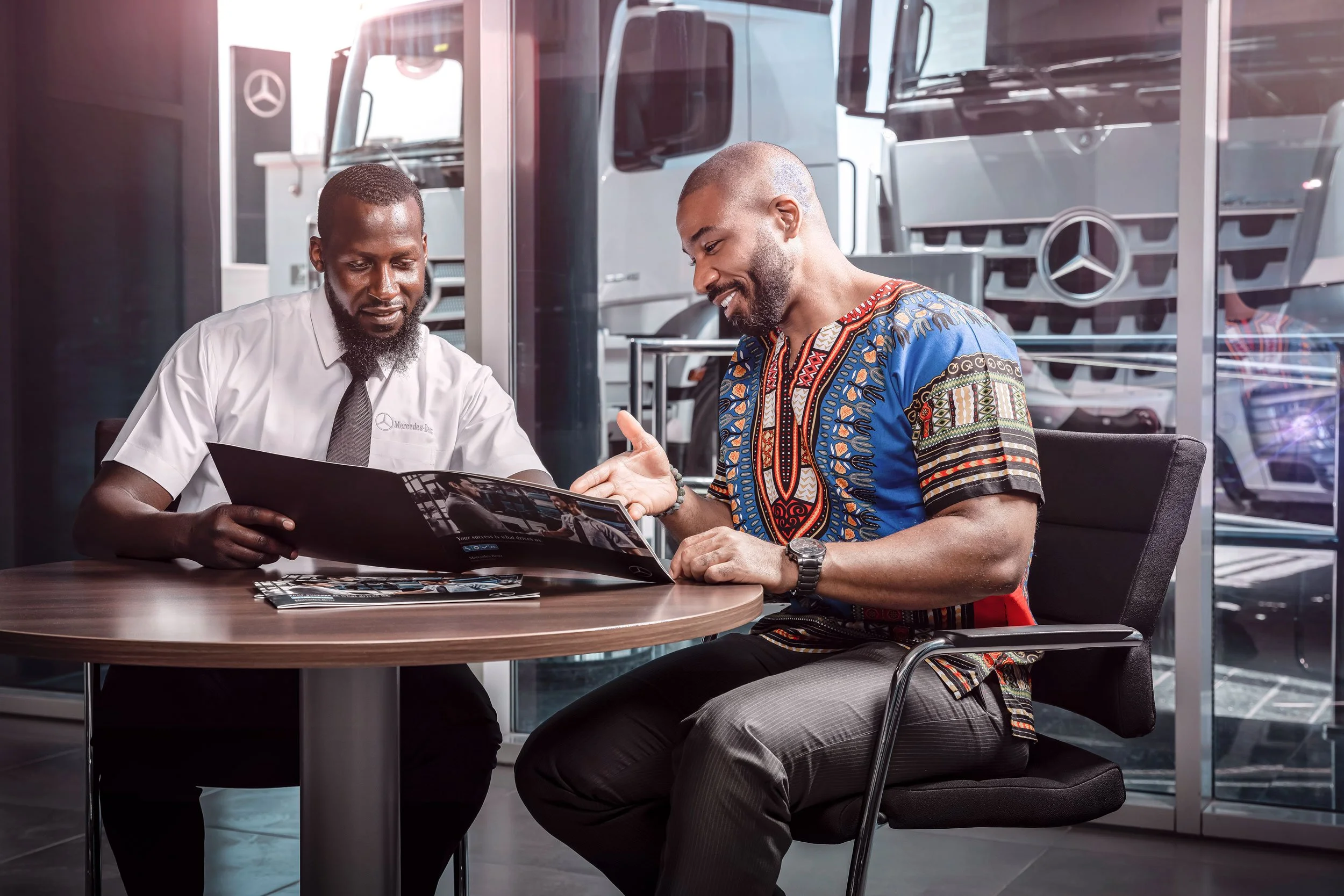 A man in a colorful shirt sits at a table with a salesperson in a Mercedes-Benz dealership, looking at a brochure with large trucks visible through the glass window behind them.