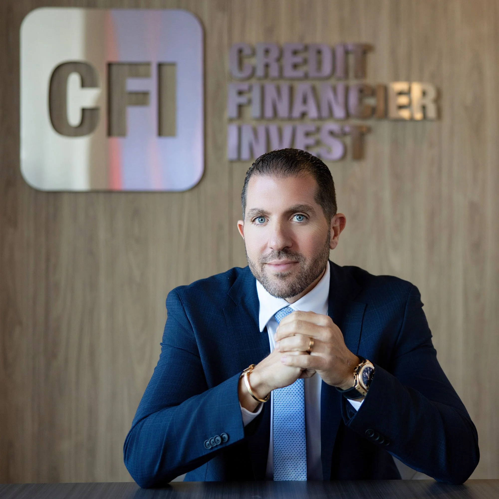 A man in a dark suit and light blue tie sitting at a desk with his hands clasped, in an office setting with wood-paneled wall and words like 'CREDIT', 'FINANCER', and 'INVEST' on the wall behind him.