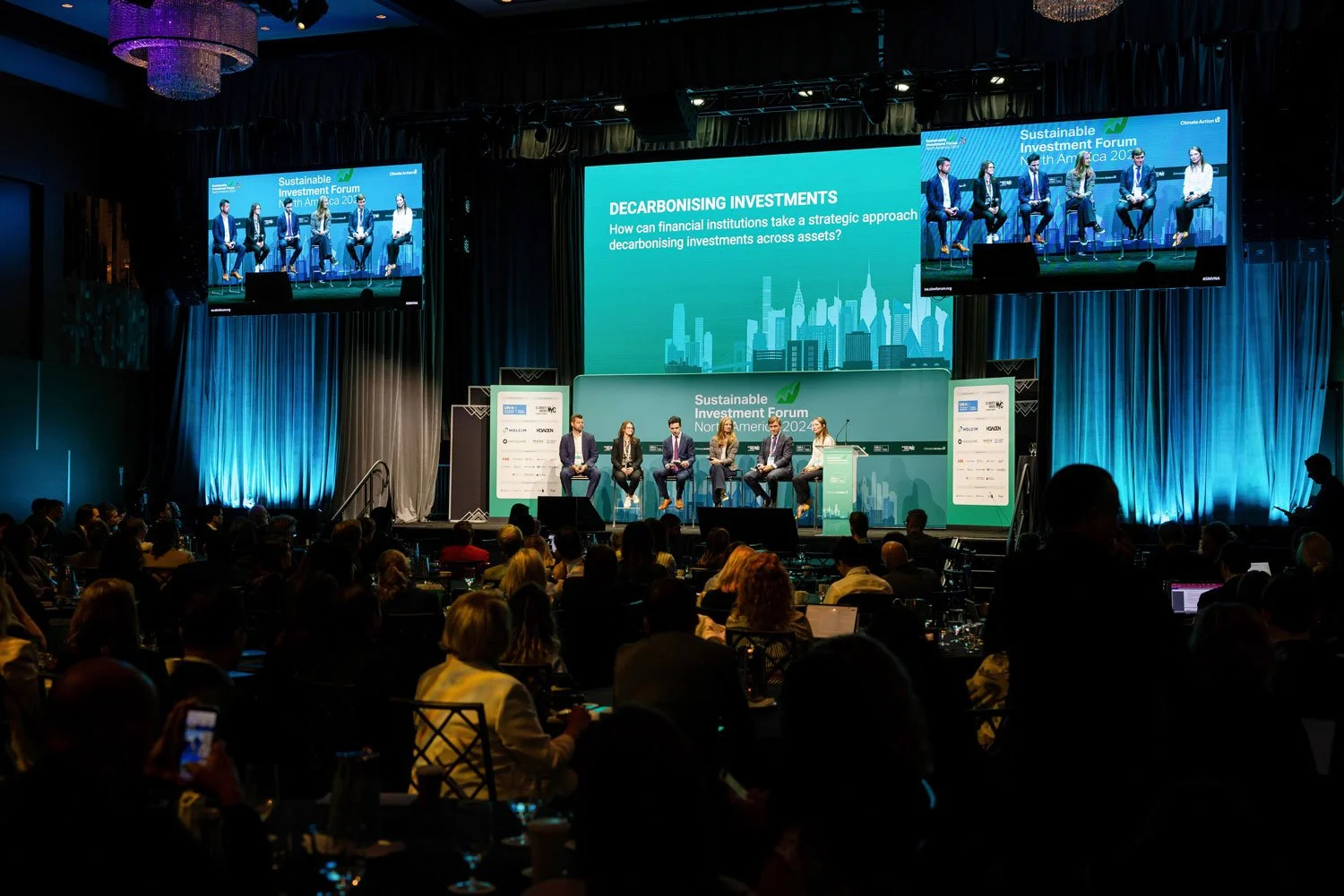 A panel discussion at the Sustainable Investment Forum North America 2024, with six speakers seated on stage under large screens displaying the forum's theme and topic on decarbonizing investments, set in a dimly lit conference hall filled with an au
