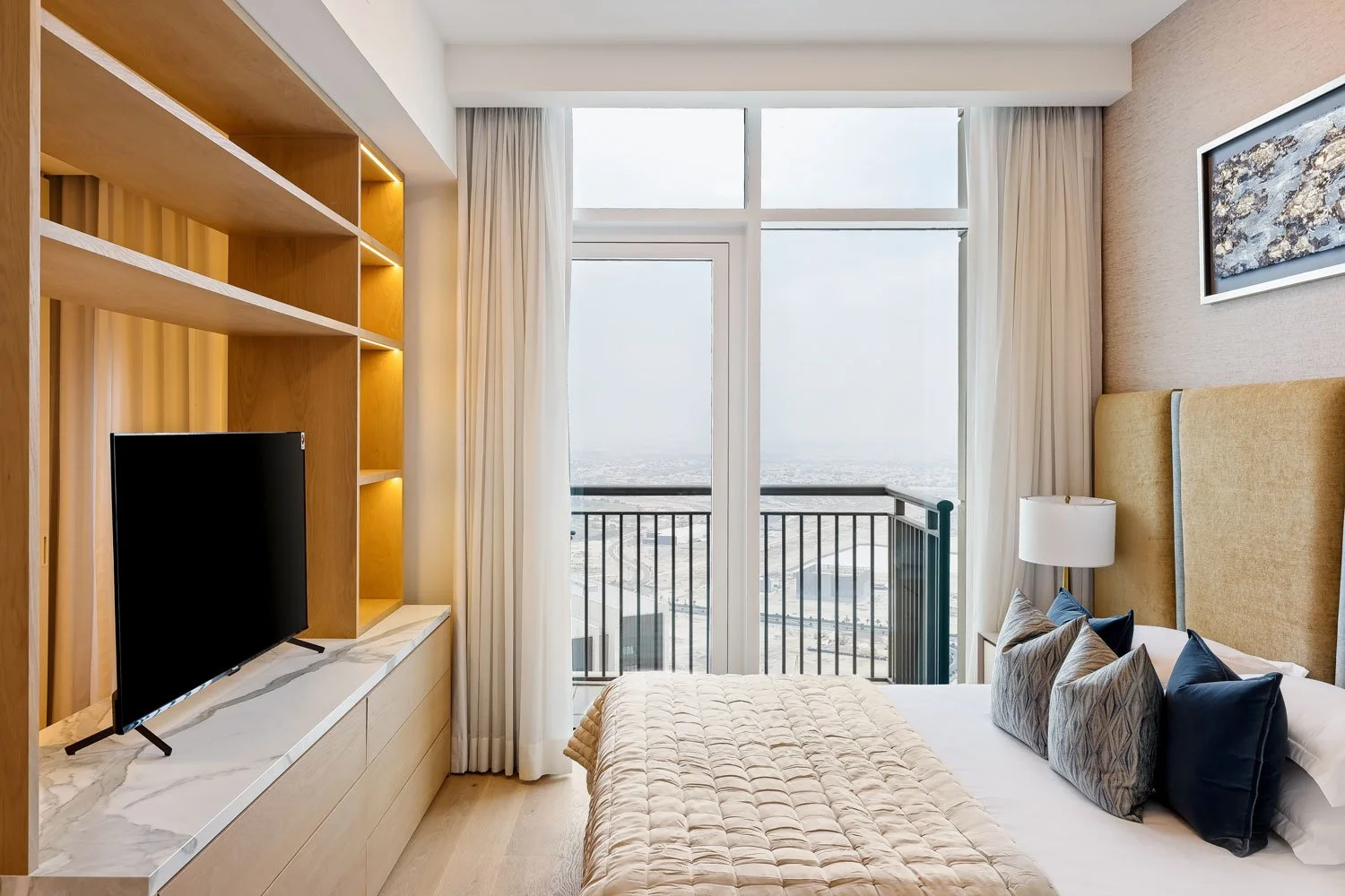 A modern hotel room with a large window overlooking a cityscape, a bed with multiple pillows, a bedside lamp, a wooden shelf with a television, and beige and white decor.
