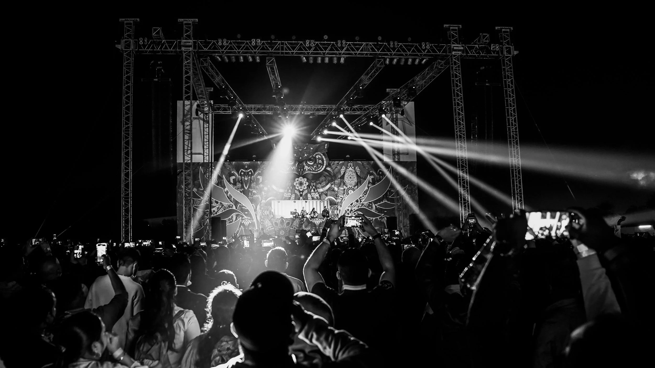 Black and white photo of a lively crowd at a concert or music festival, with a stage illuminated by bright lights and electronic equipment, and an elaborate backdrop featuring artistic designs.