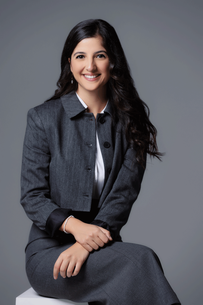 A woman with long dark wavy hair smiling, wearing a gray business suit, sitting on a white block against a gray background.