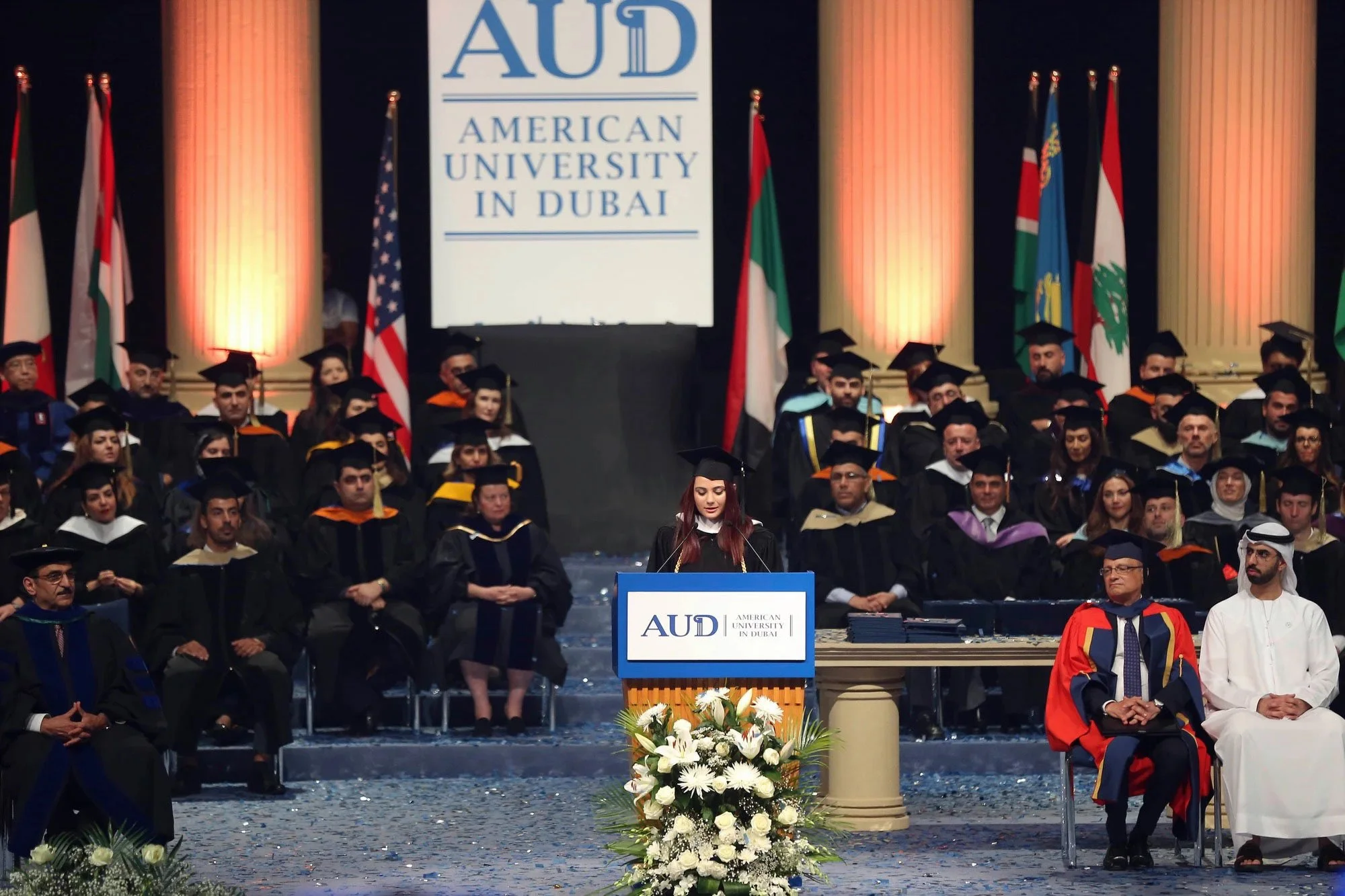 A graduation ceremony at American University in Dubai, with graduates in caps and gowns seated on stage, a woman speaking at a podium decorated with flowers, and flags from various countries displayed behind.