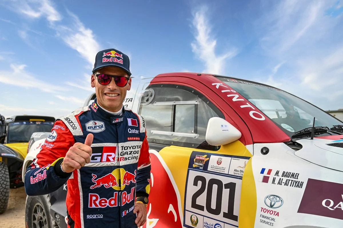 Motorsport driver standing beside rally vehicle during Dakar race event coverage.