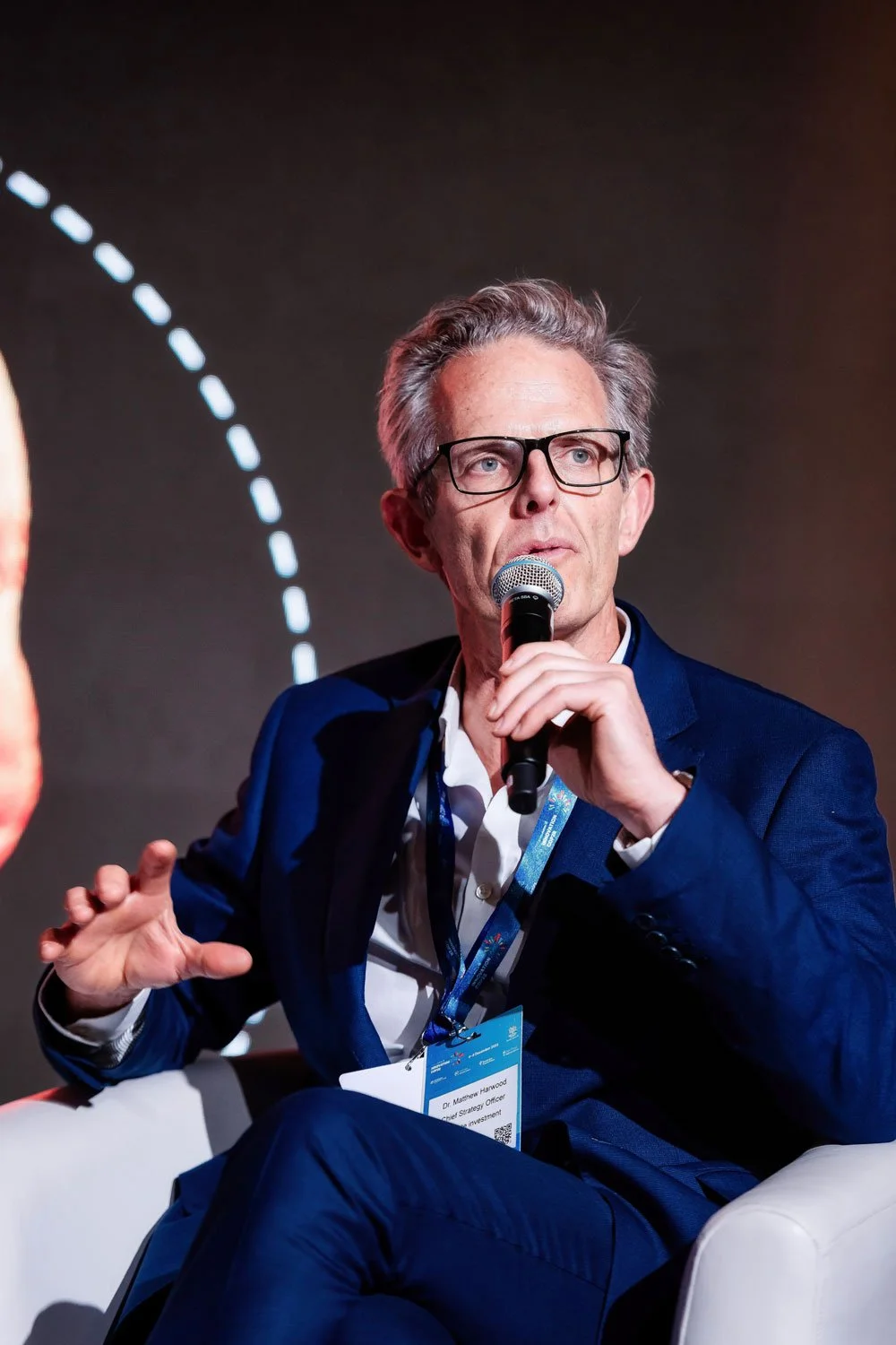 An older man with gray hair and glasses speaking into a microphone, wearing a blue suit and sitting on a white chair.