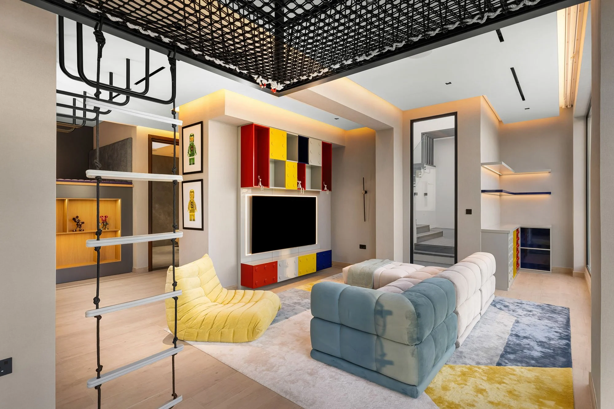Modern living room with a pastel sectional sofa, a yellow lounge chair, and a black entertainment center with a large flat-screen TV, colorful storage units, and decorative Lego artwork on the wall.