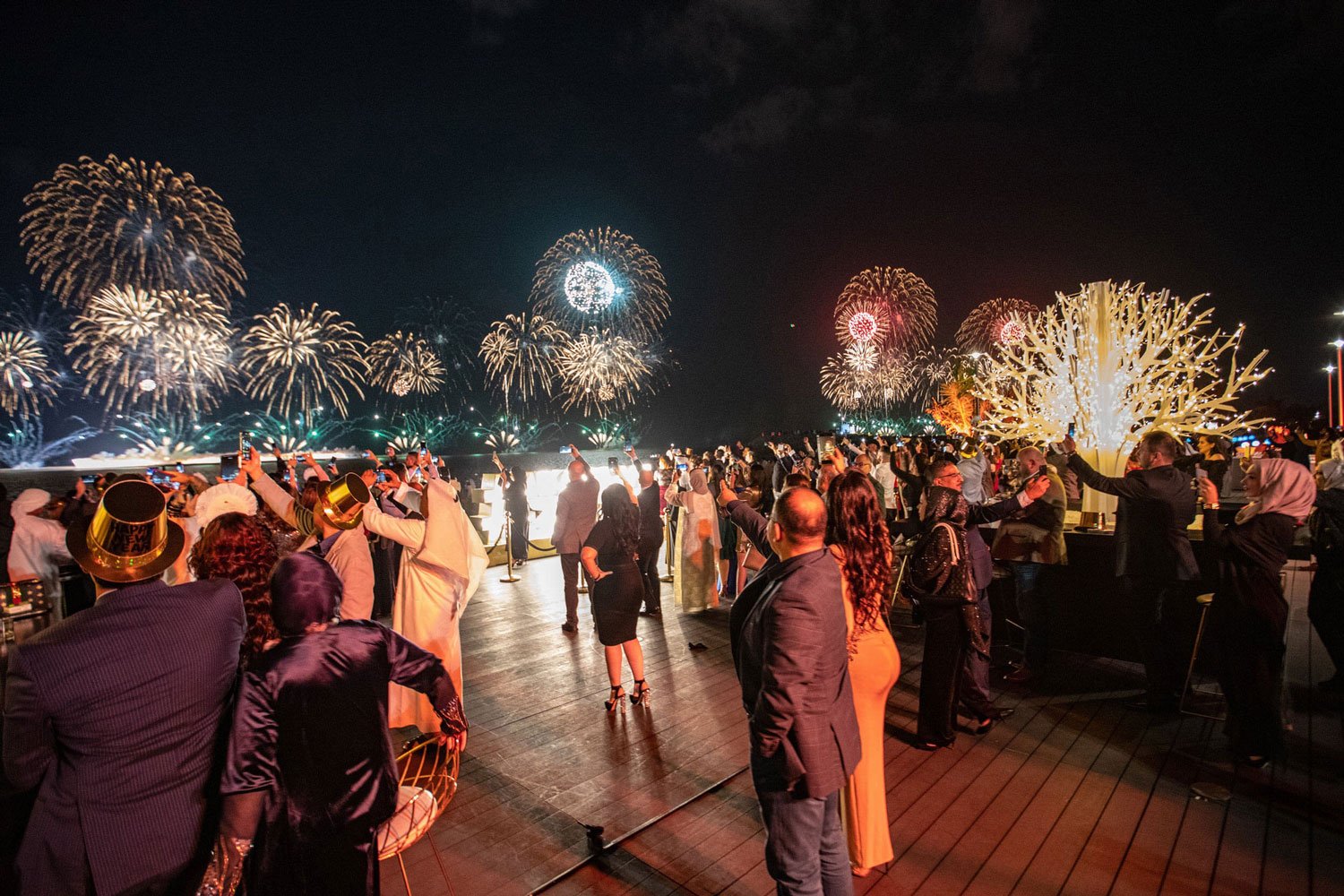 Nighttime celebration on a rooftop with people watching fireworks display in the sky. Some are taking photos or videos, and all are dressed in formal attire. The scene includes a decorative white sculpture resembling coral and a bright, colorful fire