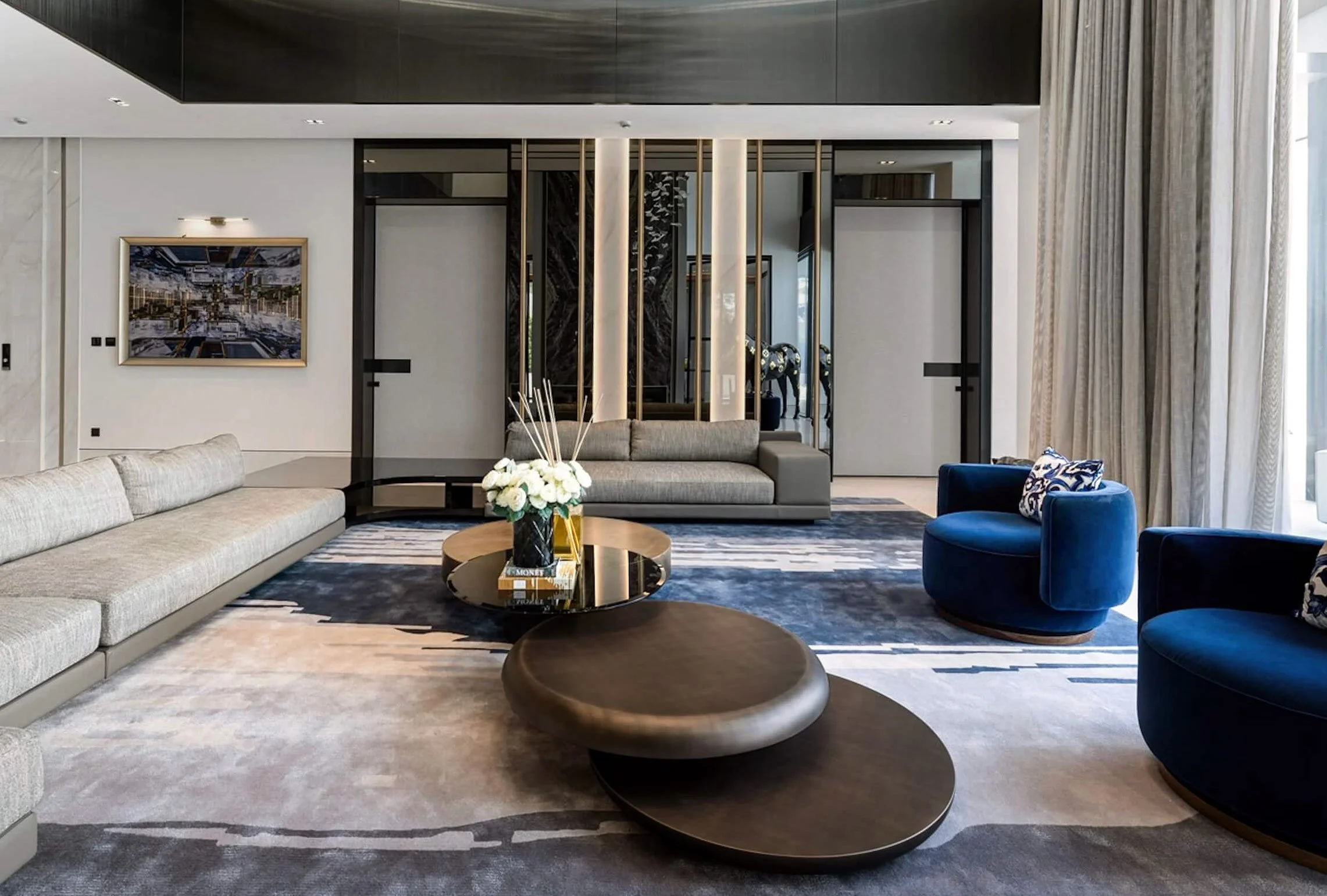 Modern hotel lobby with gray and blue seating, a black circular coffee table with a flower arrangement, abstract area rug, large curtains, and contemporary wall art.