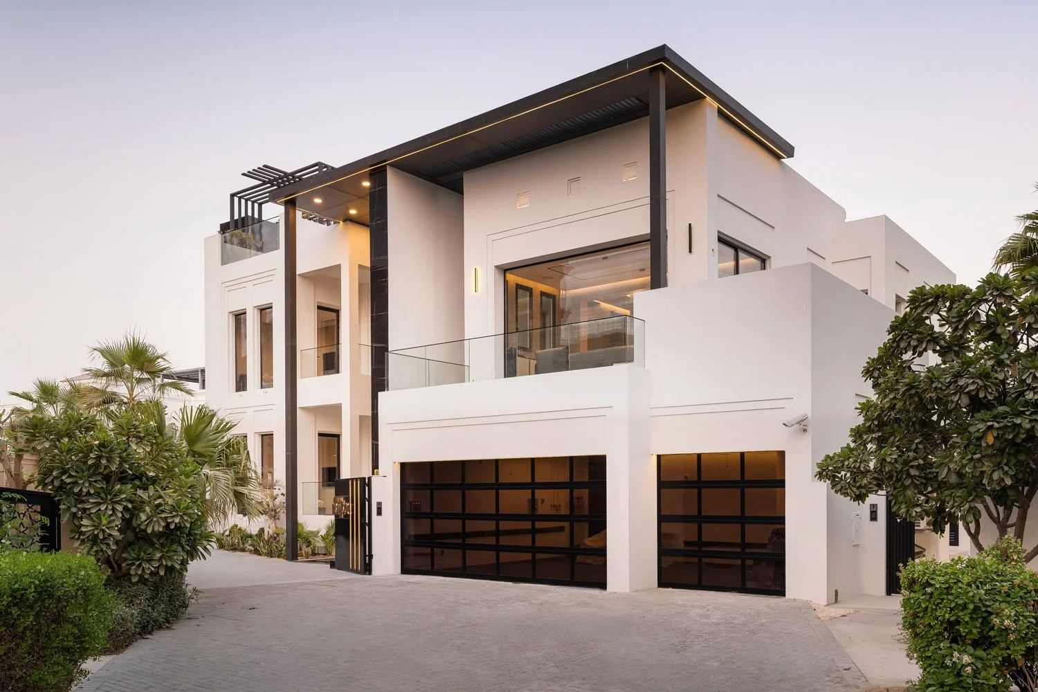 Modern white multi-story house with black accents, large glass windows, a garage with black doors, surrounded by greenery, during dusk