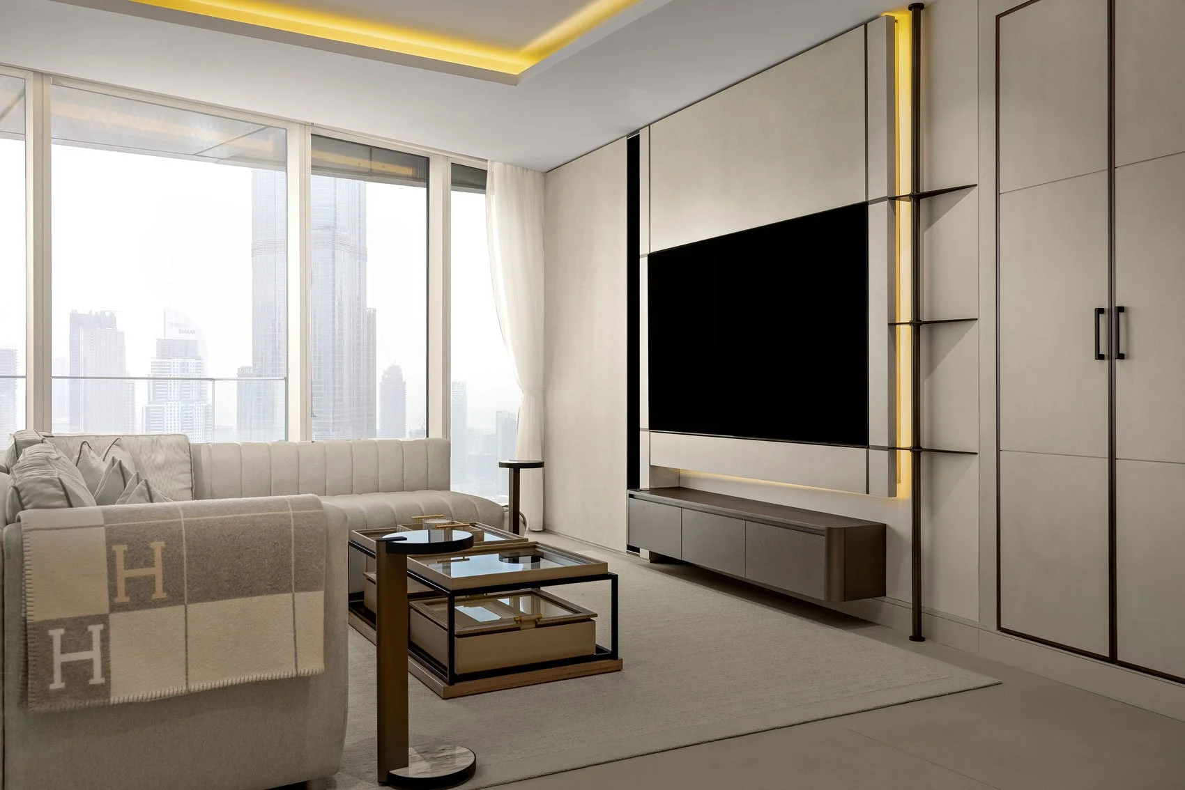 Modern living room with large windows, a white sofa with cushions, a black TV mounted on a wall with built-in cabinets and shelves, a beige area rug, and city skyline view in the background.