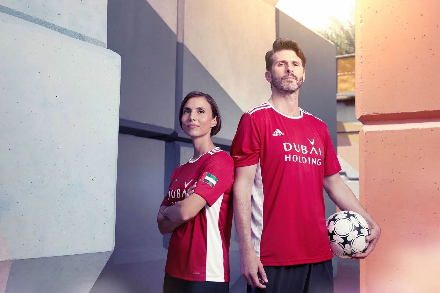 A woman and a man in red soccer jerseys standing outdoors among large geometric concrete blocks, with the man holding a soccer ball.