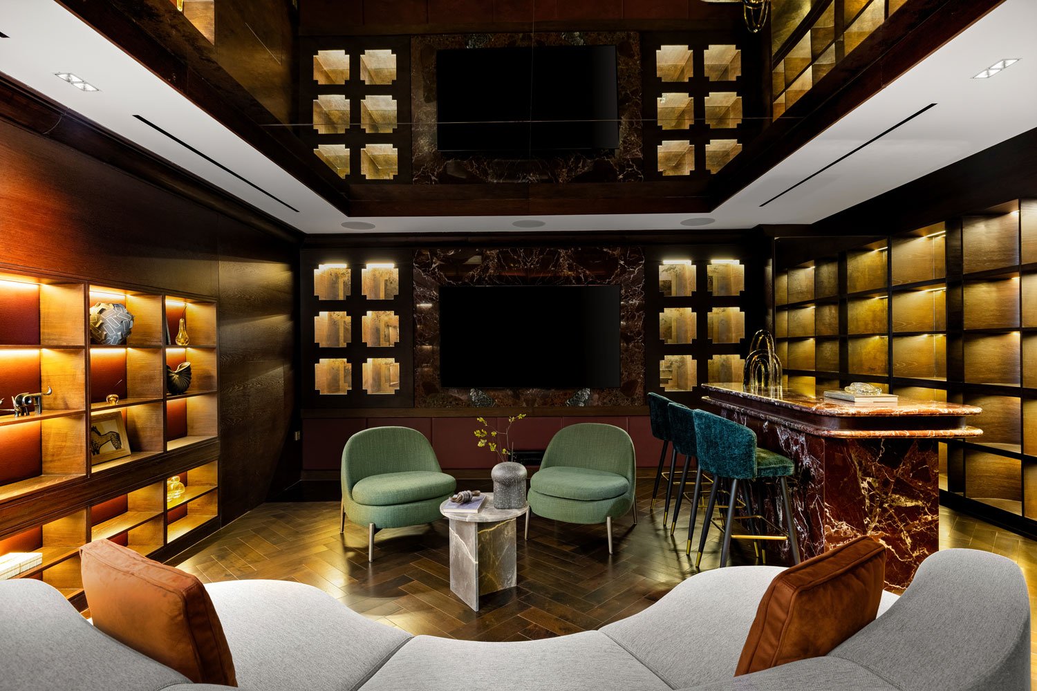 A cozy living room with dark wood paneled walls and gold accents. There are built-in shelves with decorative objects, a large flat-screen TV mounted on a marble wall, and a bar area with a marble counter. The seating includes a gray sofa with brown p