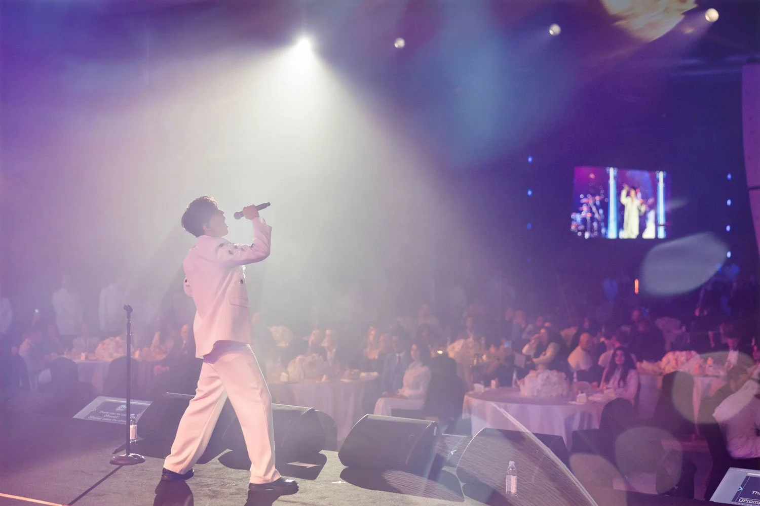 Performer singing with a microphone on stage at a concert, with audience seated at tables in the background and stage lights shining down.