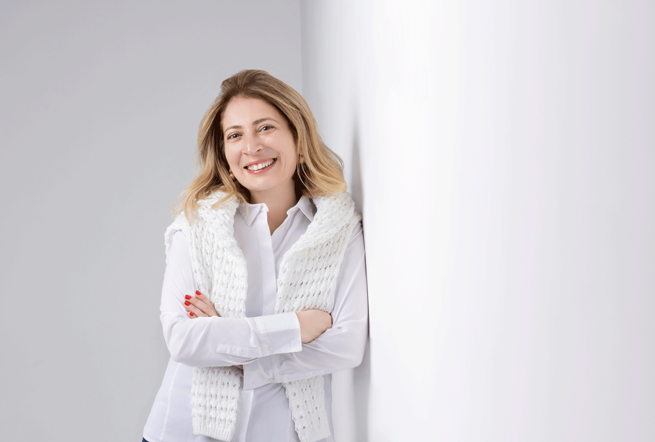 A woman with shoulder-length blonde hair wearing a white shirt and a white knitted scarf, smiling and standing with arms crossed against a light gray wall.
