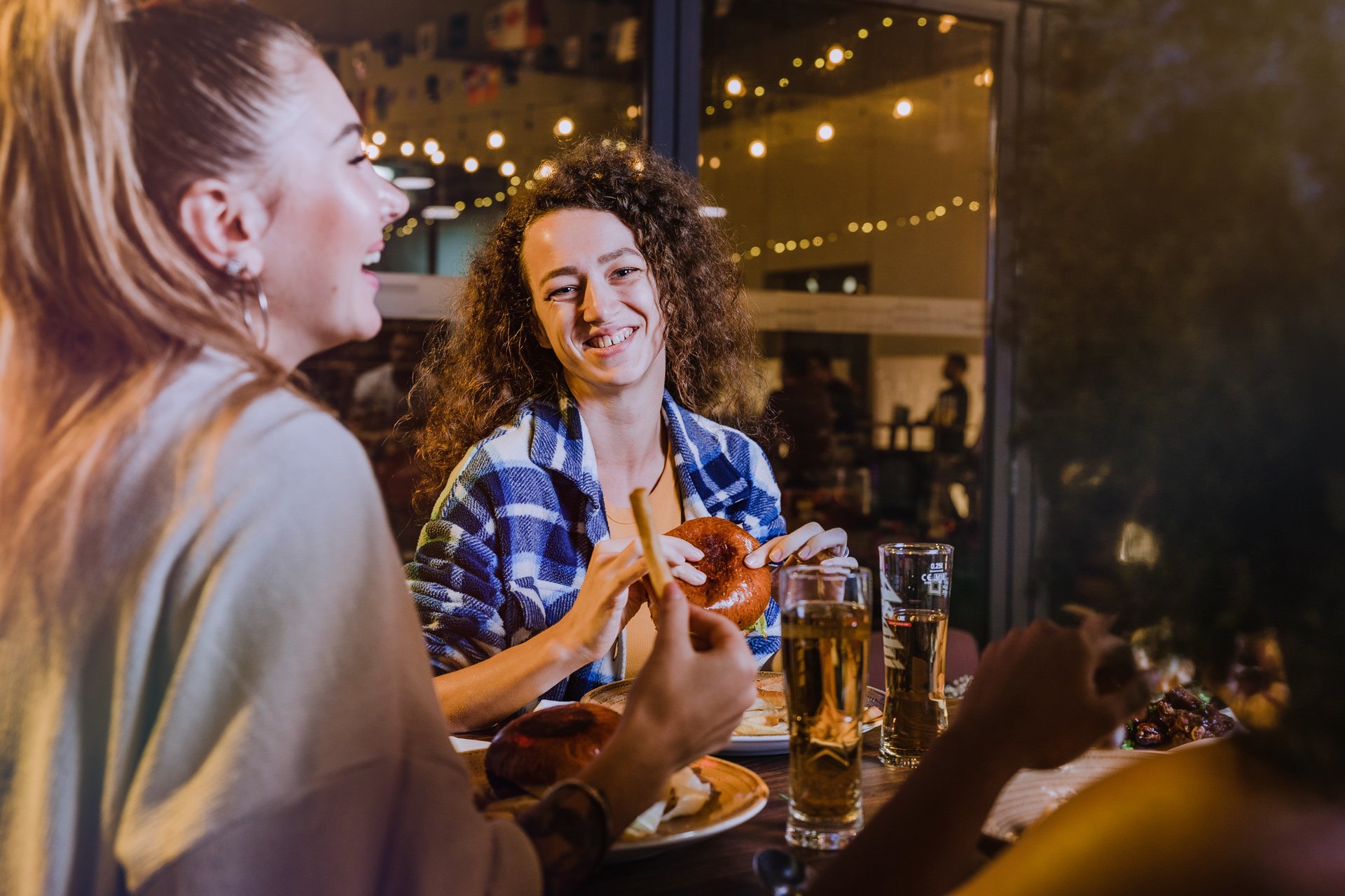 Three women sitting at a table enjoying food and drinks at a restaurant, with the background decorated with string lights.