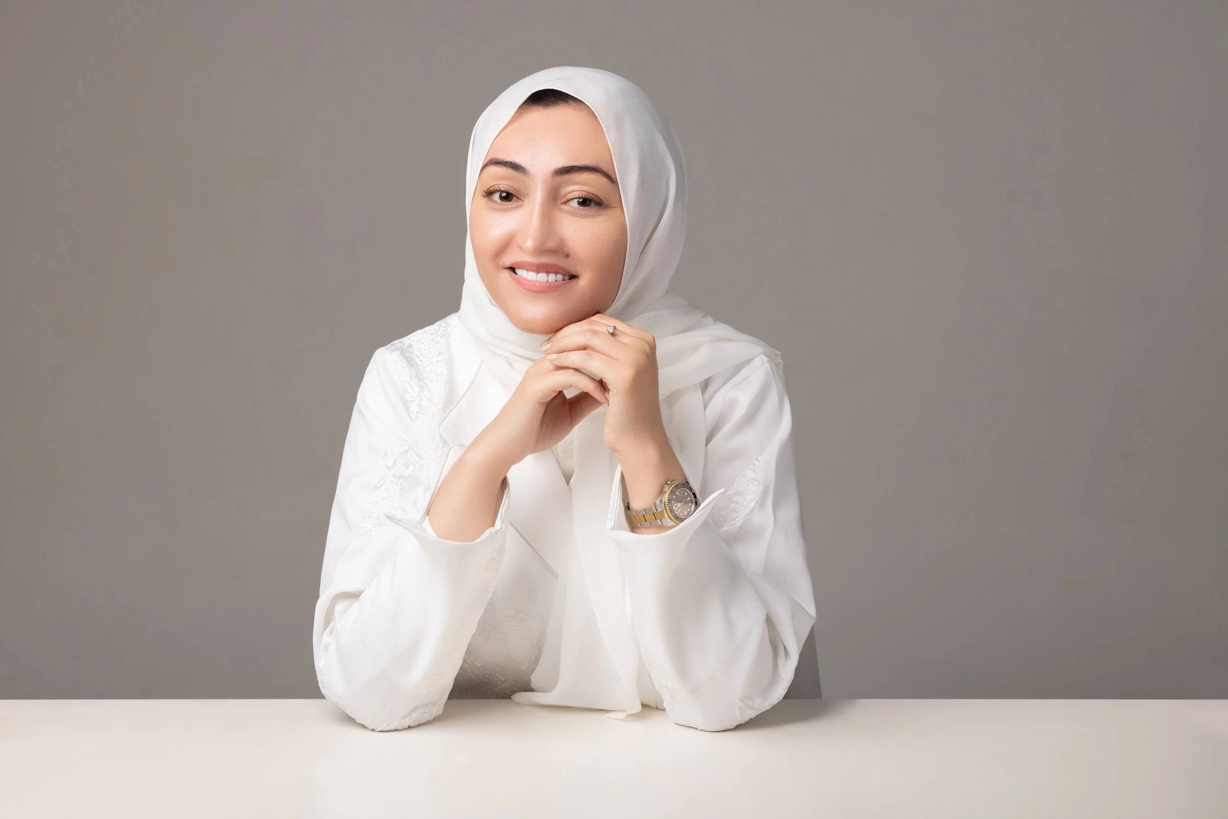 A woman wearing a white headscarf and white blouse, smiling, sitting at a table against a plain gray background.