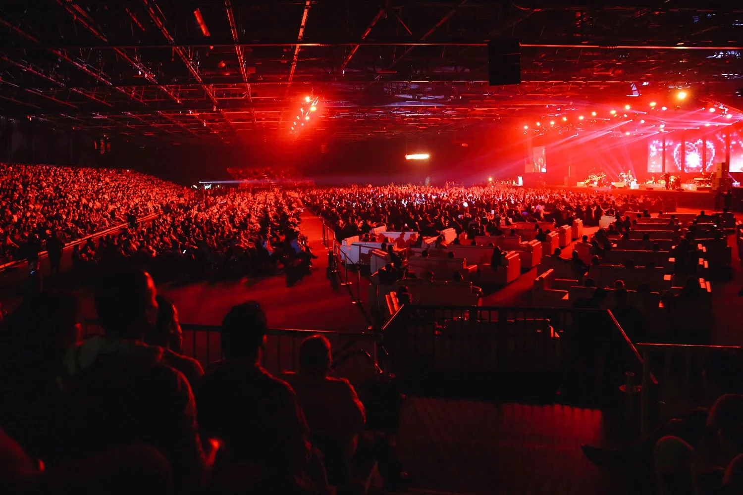 A large indoor concert venue filled with an audience seated in front of a stage illuminated in red lights.