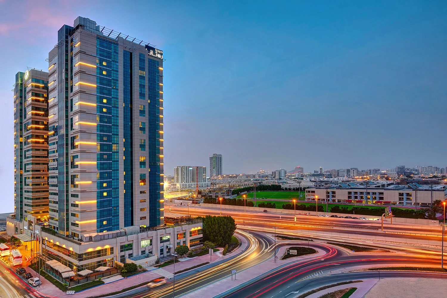Cityscape with tall modern buildings and light trails from moving vehicles on curved roads at dusk.
