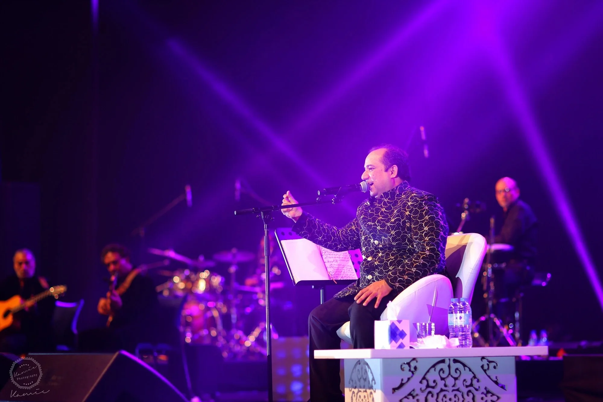Performer singing into a microphone on stage, dressed in a patterned jacket, with musicians playing guitar and drums in the background, illuminated by purple stage lights.