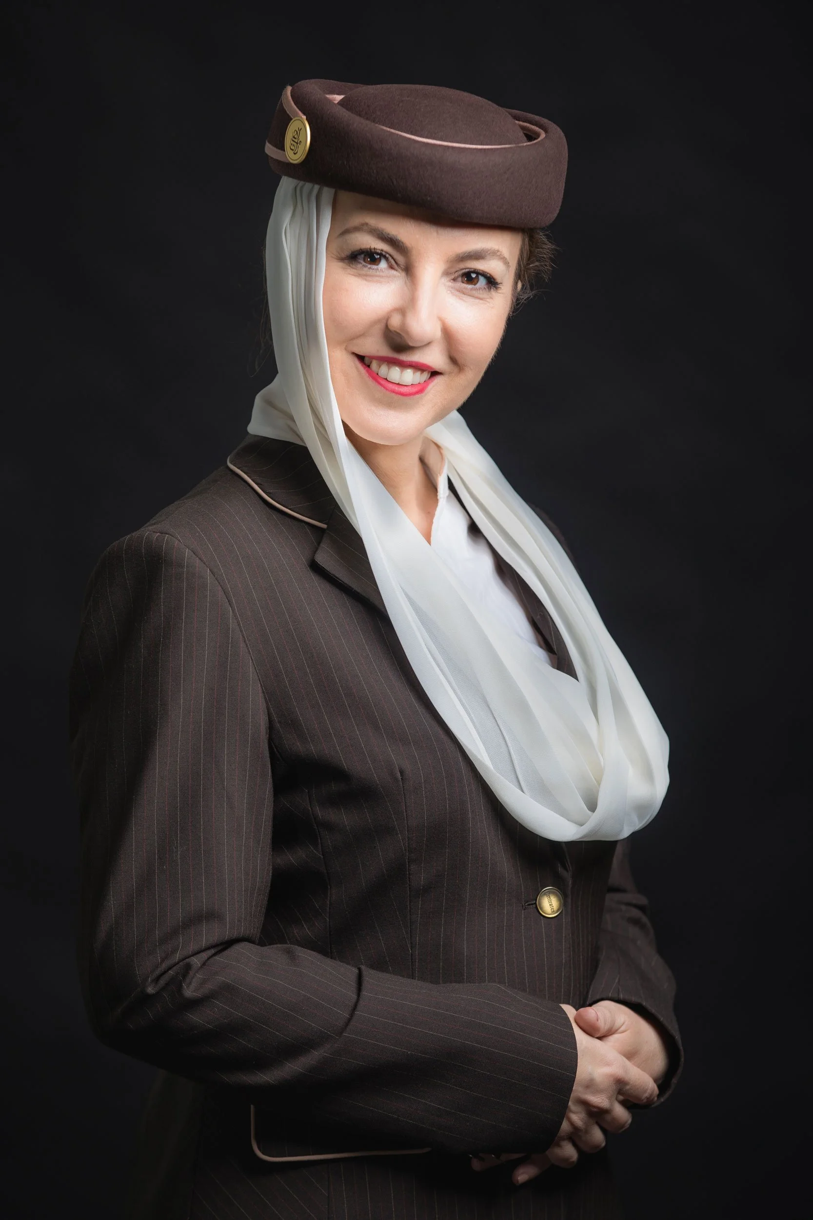 A woman smiling, wearing a vintage-style brown hat with a pin, a white scarf, and a brown pinstripe suit against a black background.