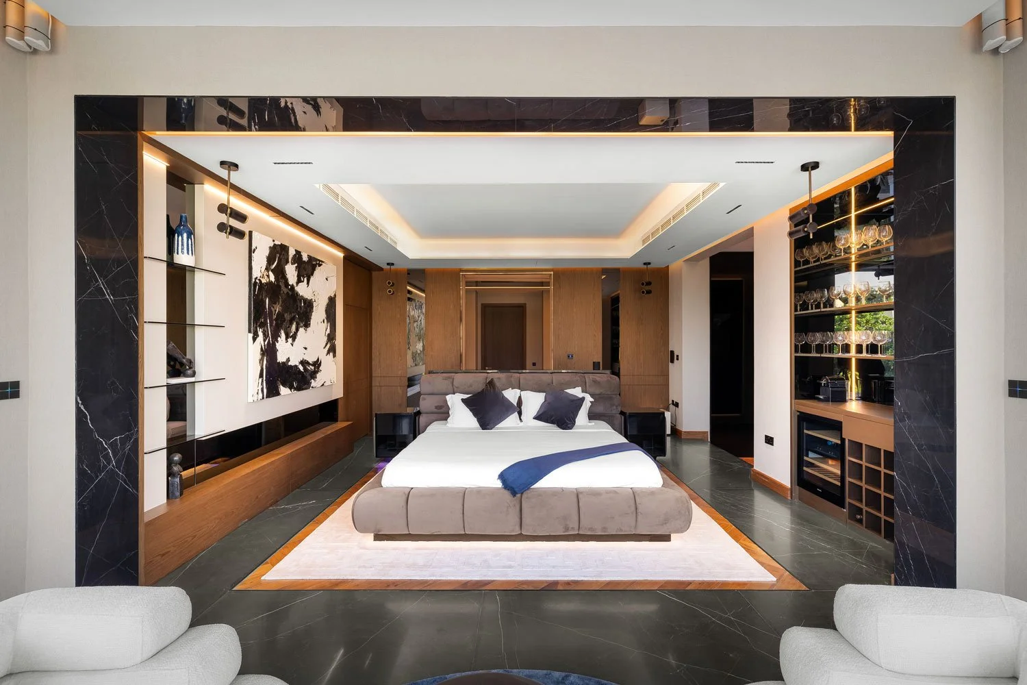 A modern hotel room with a king-size bed, black pillows, a beige headboard, and a blue throw blanket. The room has black marble flooring, wood paneling, and built-in shelves with glassware on the right. There is seating in the foreground and artistic