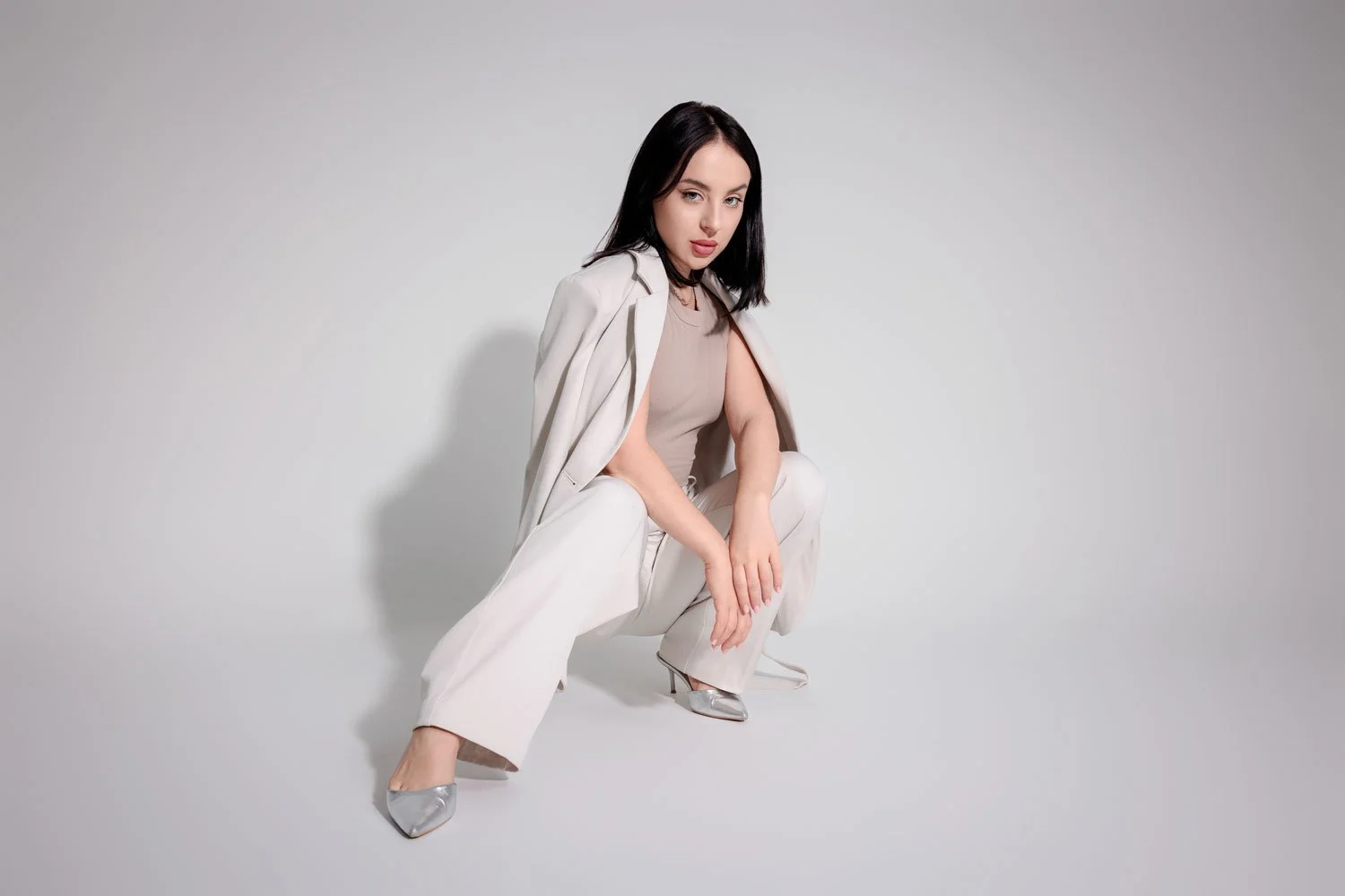 A woman with black hair, dressed in a beige top and white pants, squatting against a plain white background.