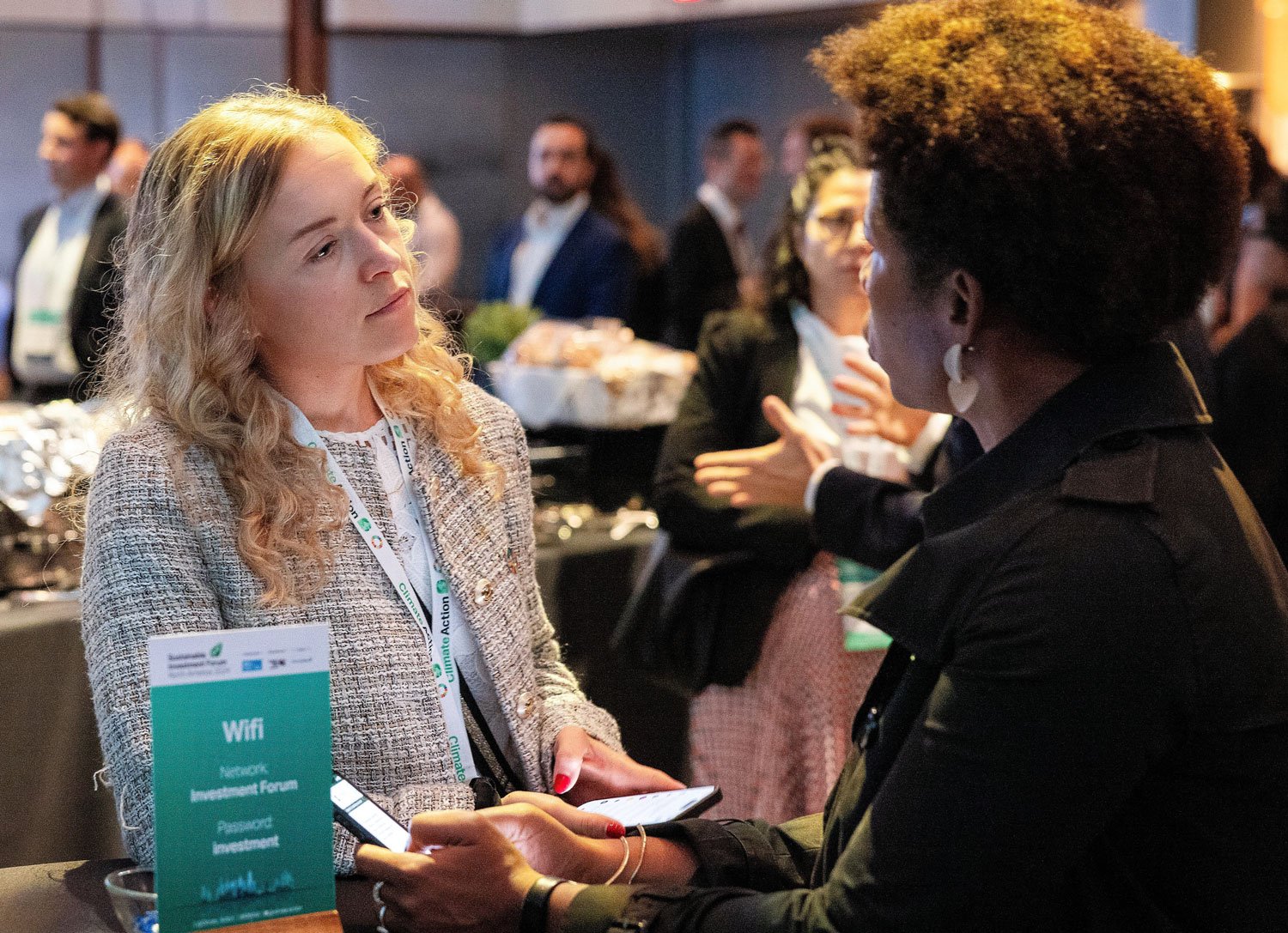 Two women engaged in a conversation at a professional event, with blurred people and a table with refreshments in the background.