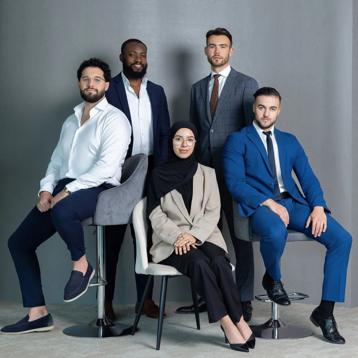 Six professionally dressed diverse individuals posing against a neutral gray wall, with some seated on chairs and others standing.