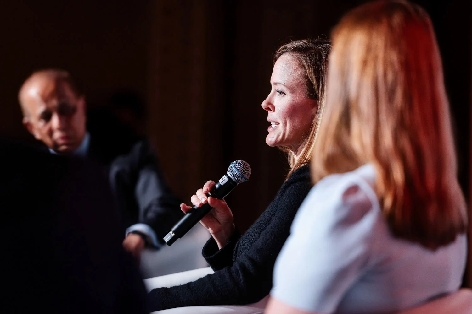 A woman speaking into a microphone at a panel discussion, with three other people partially visible, including a woman with red hair and a man in the background.