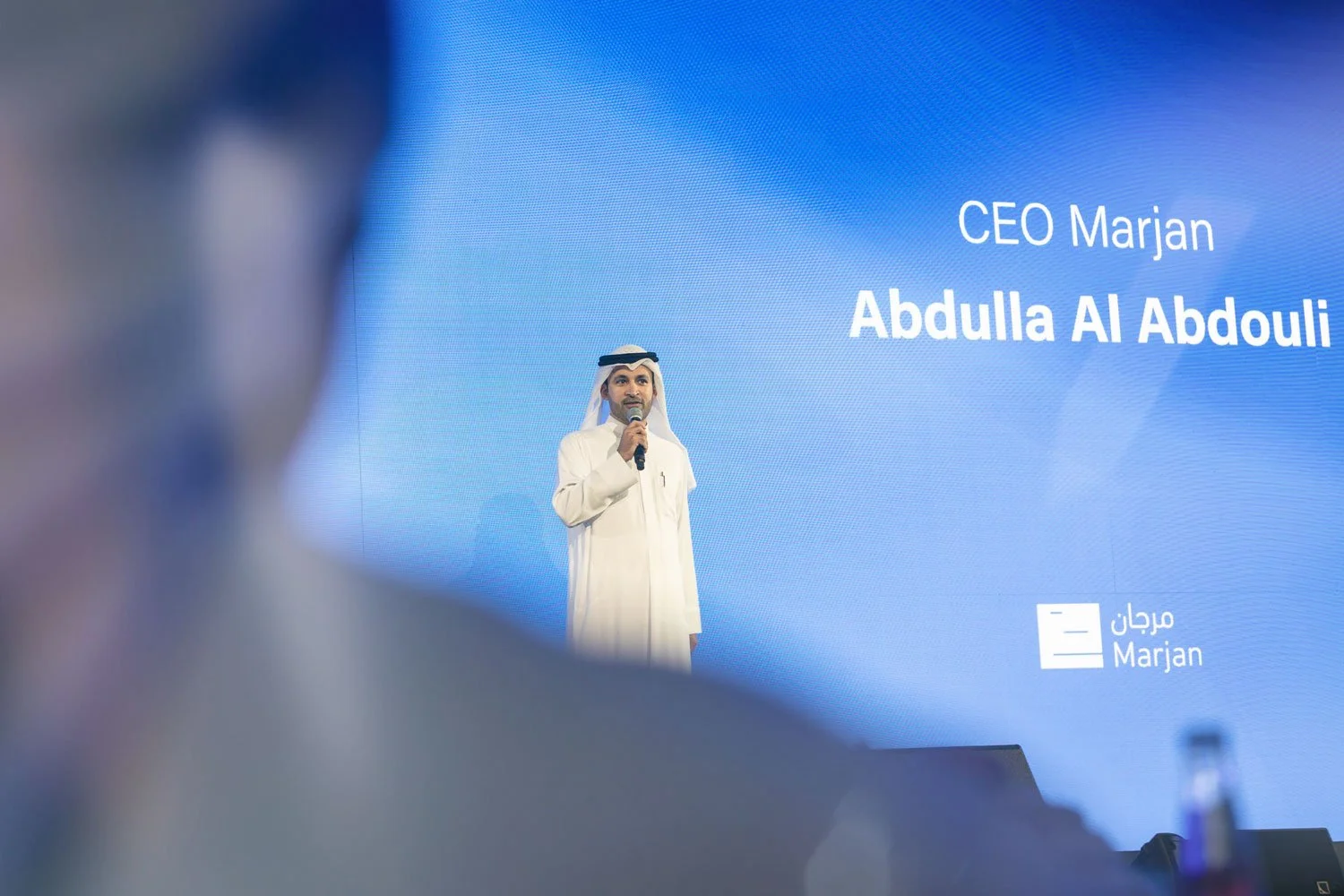 A man in traditional Middle Eastern attire speaking into a microphone on stage, with a large blue screen behind him displaying his name, Abdullah Al Abdouli, and his title, CEO Marjan.
