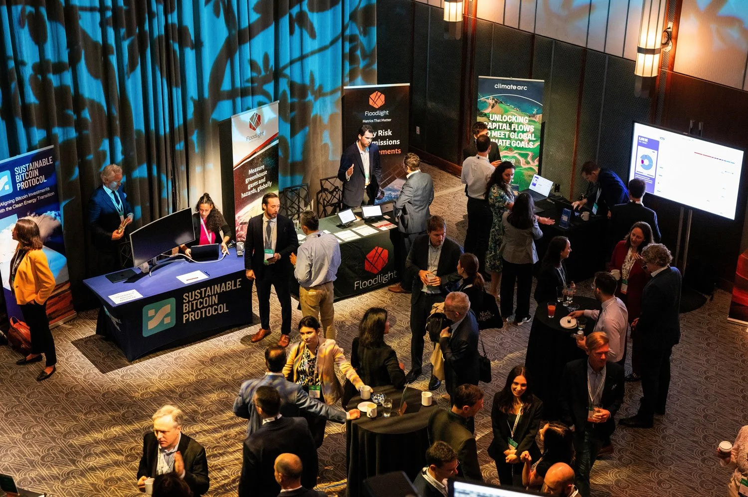 A busy conference or networking event with multiple booths and attendees engaged in conversations, some standing in groups and others sitting, with large display screens and promotional banners.