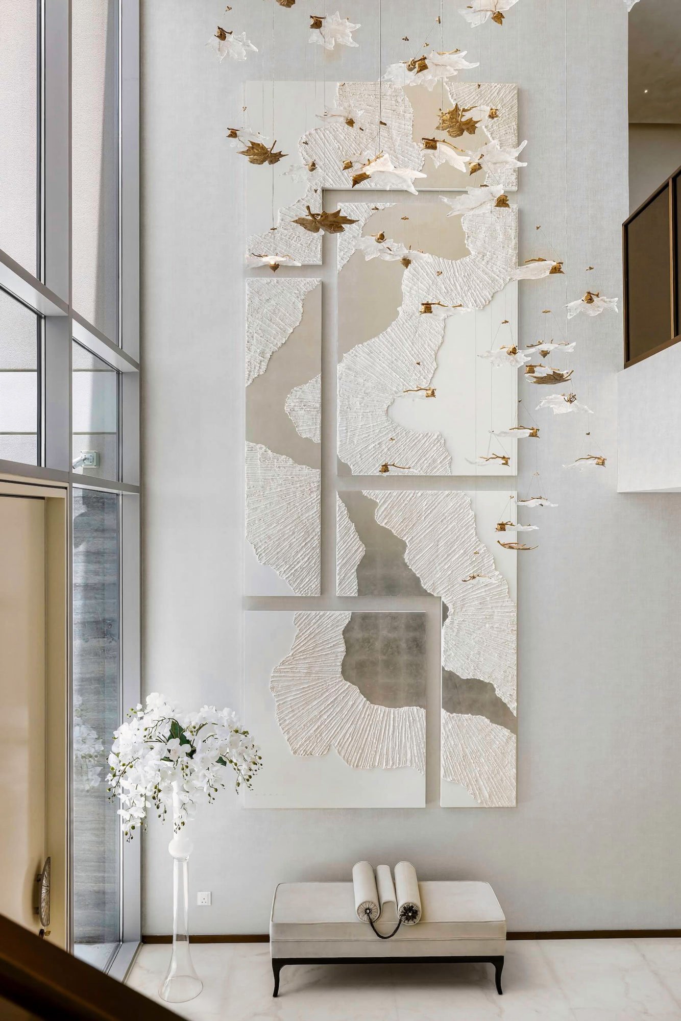 Large abstract wall art with textured white panels, featuring suspended decorative fish and waves in a modern interior with a tall flower arrangement and a bench.