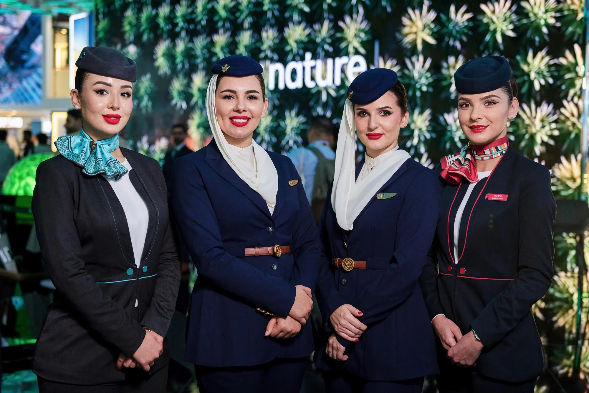 Four women in navy airline uniforms standing together at an indoor event, with a green and white floral backdrop and the word 'nature' visible in the background.
