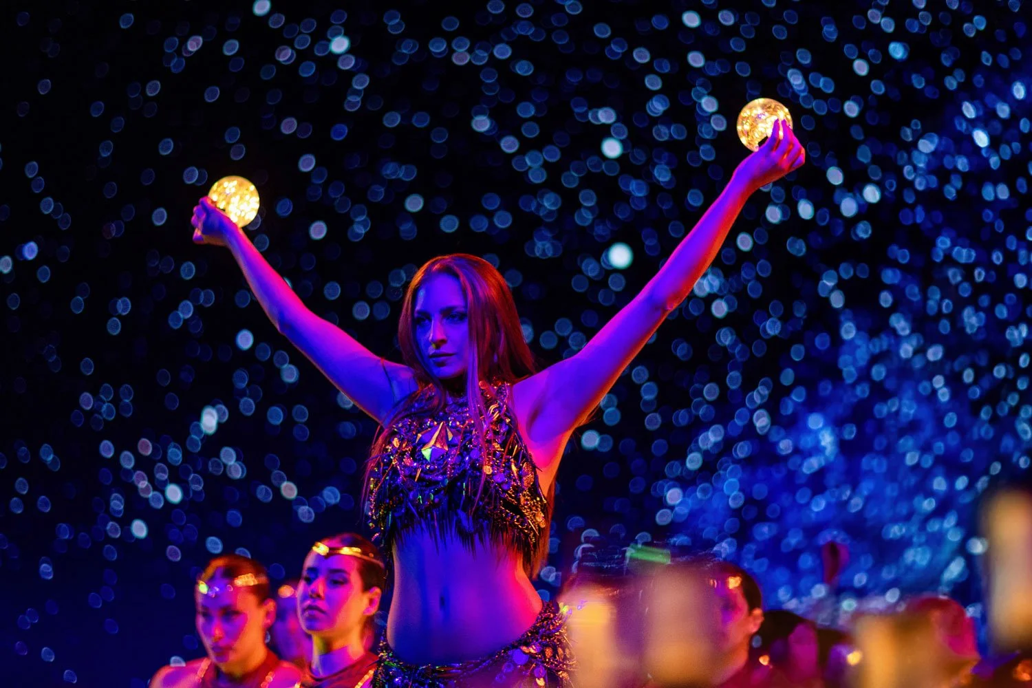 A woman with red hair in a performance outfit holding glowing orbs with arms outstretched at a night event with blue bokeh lights.