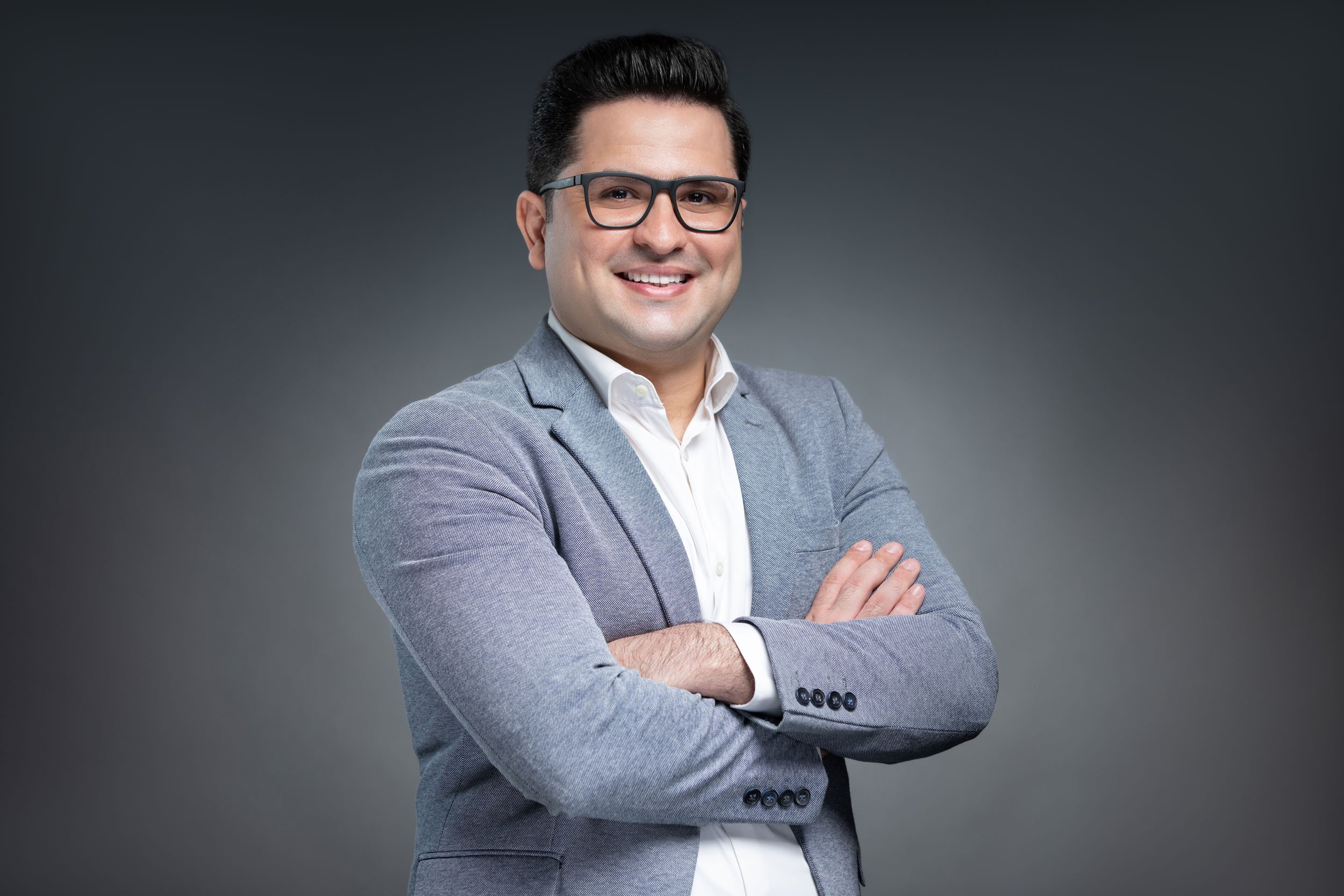 A man with dark hair, wearing glasses, a white shirt, and a gray blazer, smiling with arms crossed against a gray background.