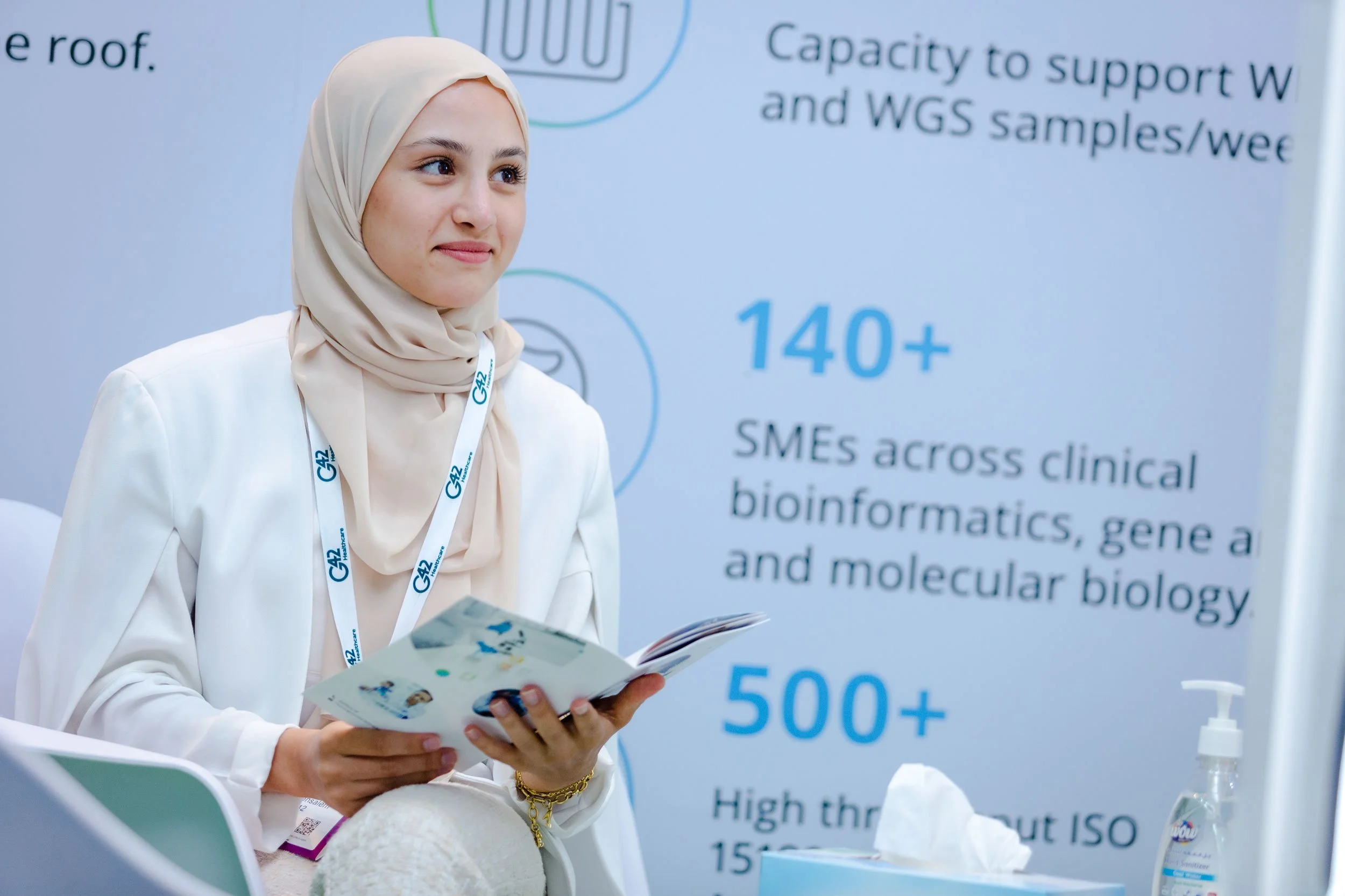 A woman wearing a beige hijab and white blazer sitting and holding a brochure at a conference or exhibition with a large infographic poster in the background highlighting bioinformatics statistics and information.