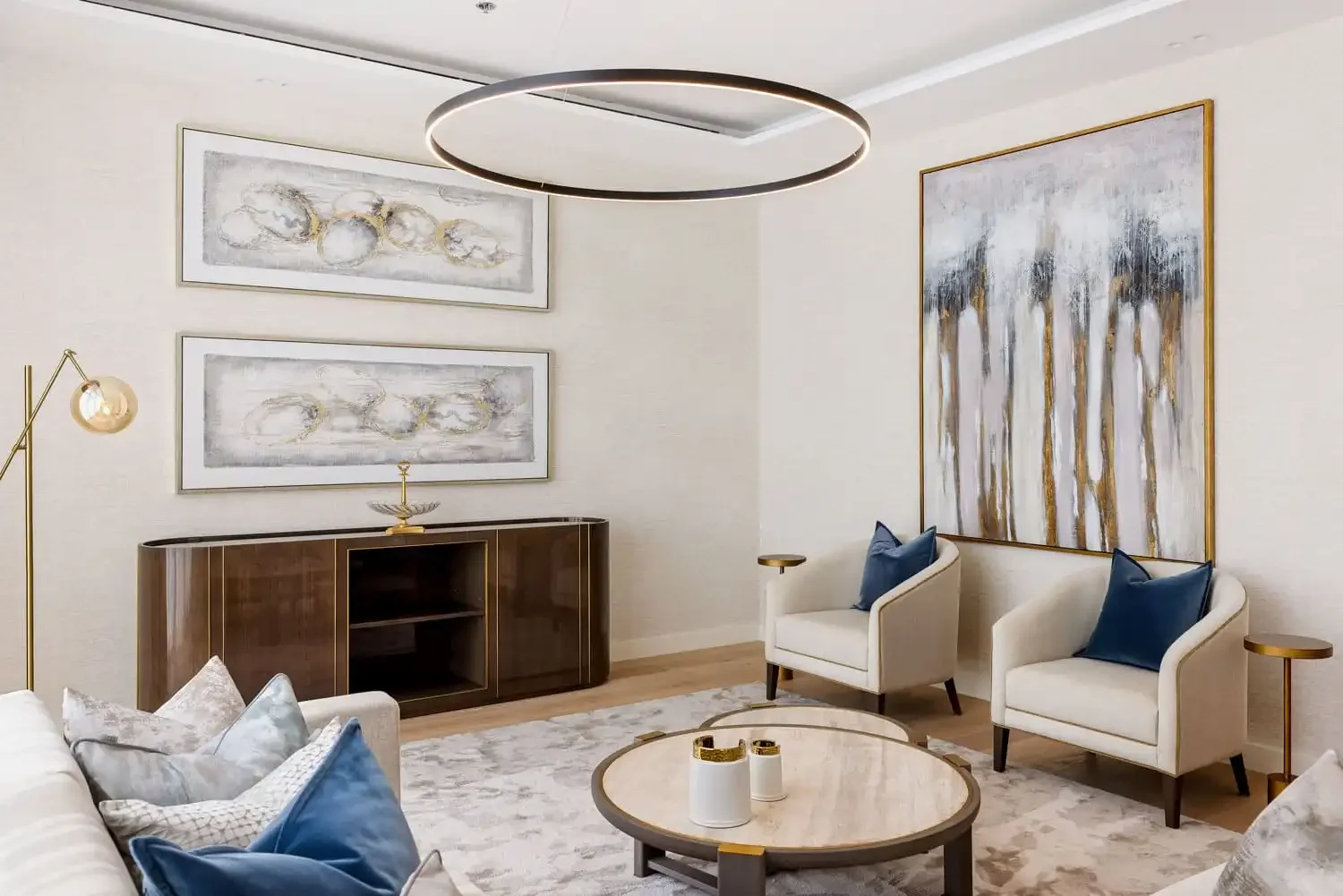 Elegant living room with white walls, abstract artwork, a round ceiling light, a beige armchair with a blue pillow, a wooden sideboard, and a round coffee table with decorative items.