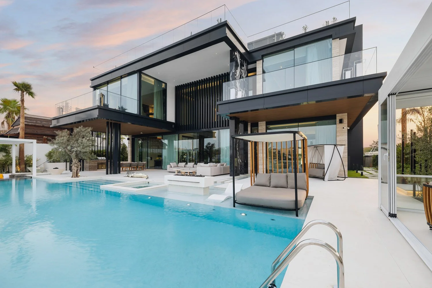 Modern living room with large windows showing a city skyline, white sofa with pillows, black coffee table with flowers, TV on wall, staircase, and dining table with place settings.
