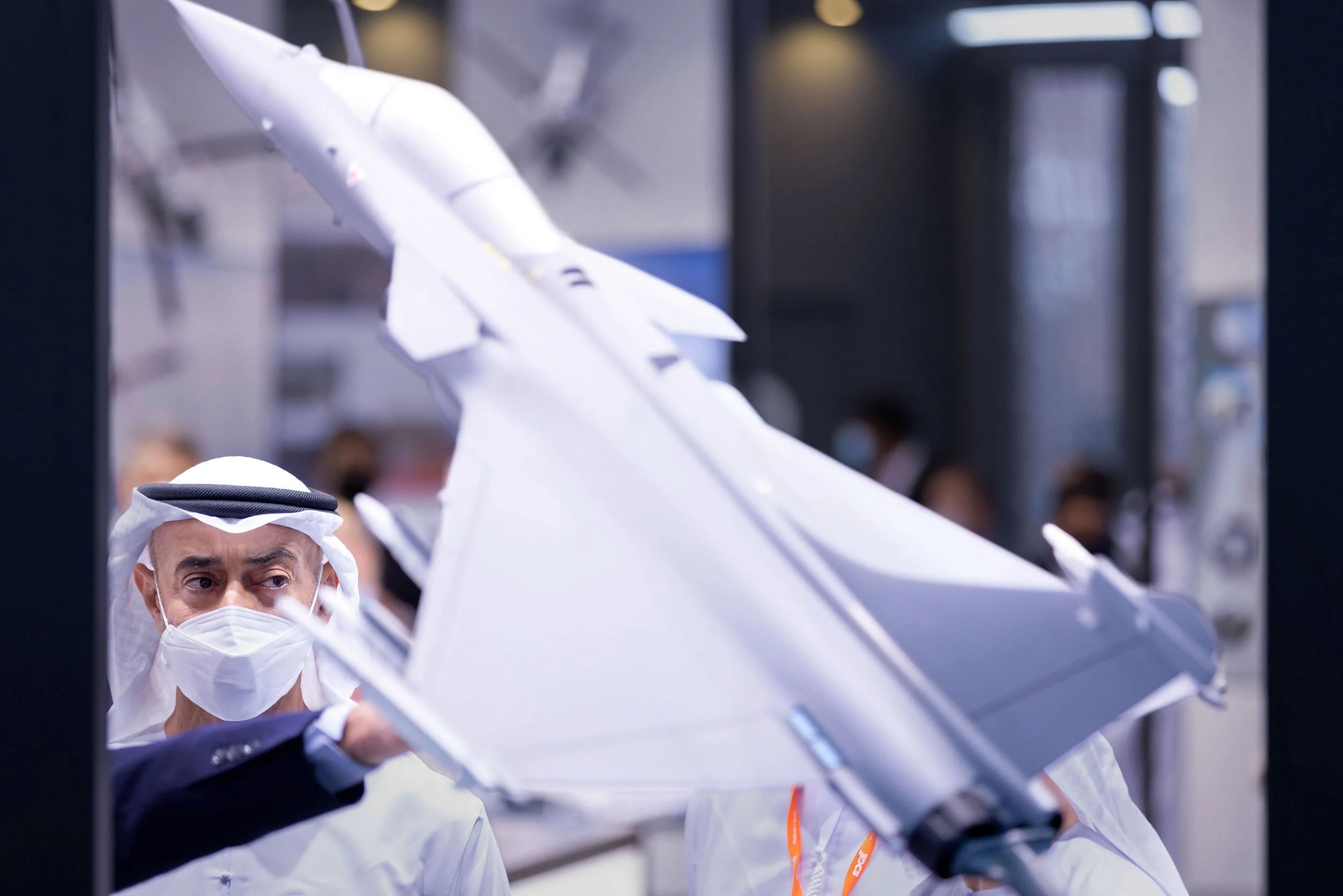 A man wearing traditional Middle Eastern attire and a face mask looking at a large model of a fighter jet.