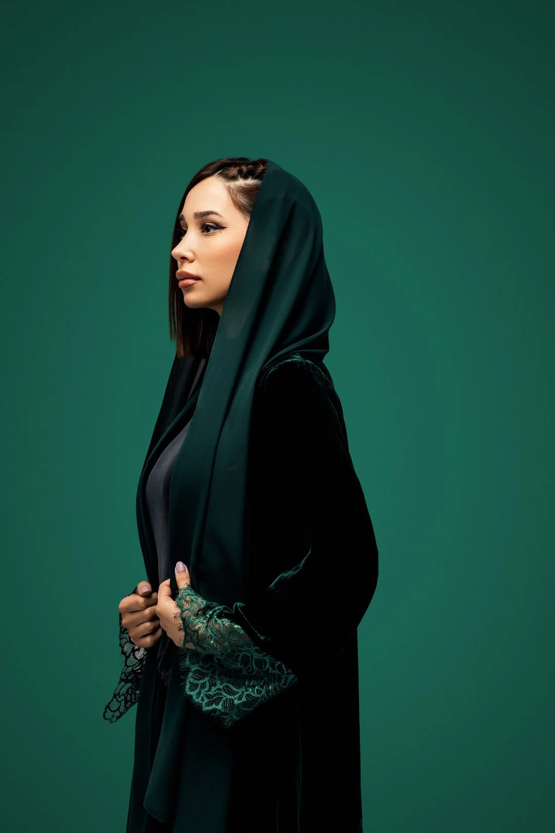 A woman with dark hair and a dark green hooded cloak with lace cuffs, standing in profile against a solid green background.
