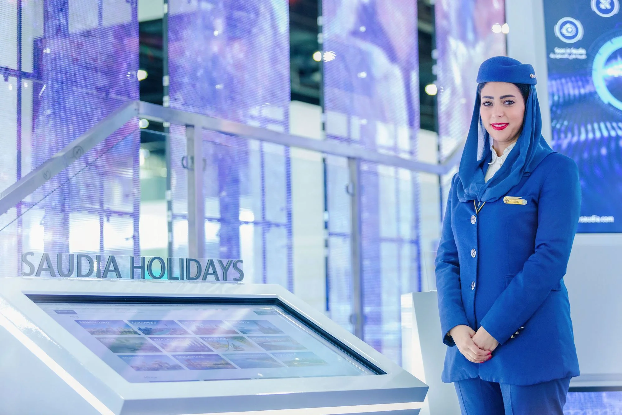 A woman in a blue uniform and matching headscarf standing next to a digital display with the text 'SAUDIA HOLIDAYS' and images of vacation destinations, inside a modern building.