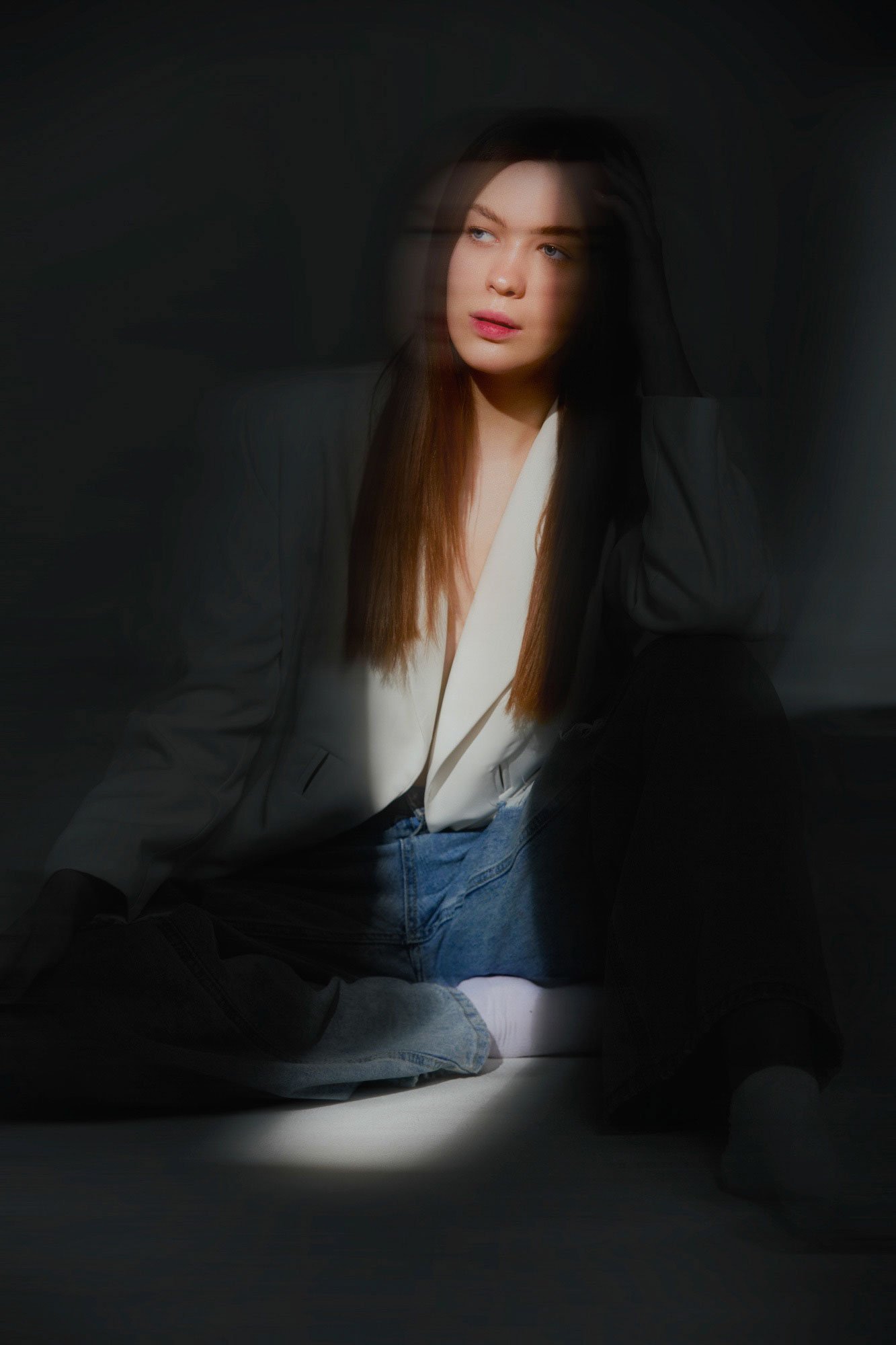 A young woman with long brown hair sitting on the floor in a dark room, illuminated by a focused light, wearing a white top, black blazer, and blue jeans, with a contemplative expression.