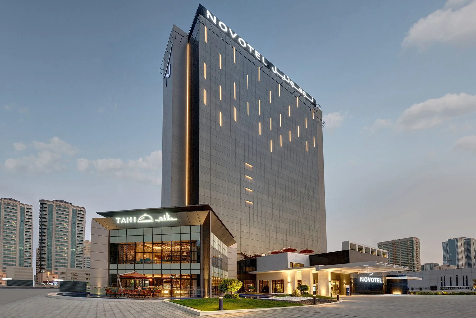 A modern tall hotel building with illuminated lights and a sign that reads 'Novotel' at the top. In front, there is a smaller building with signs in English and Arabic, including 'Tahi' and 'Novotel', with outdoor seating and a landscaped area.