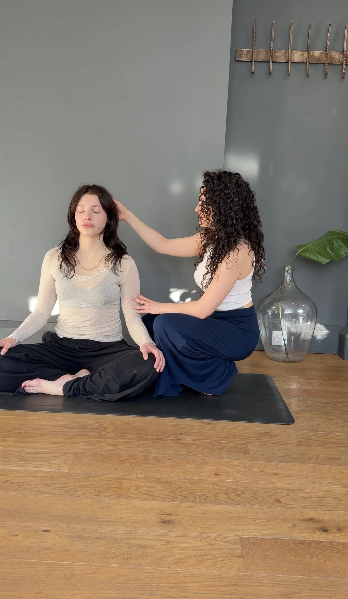 A woman with curly hair practicing energy healing or Reiki on another woman with straight hair, sitting cross-legged on a yoga mat in a room with gray walls and wooden floor.