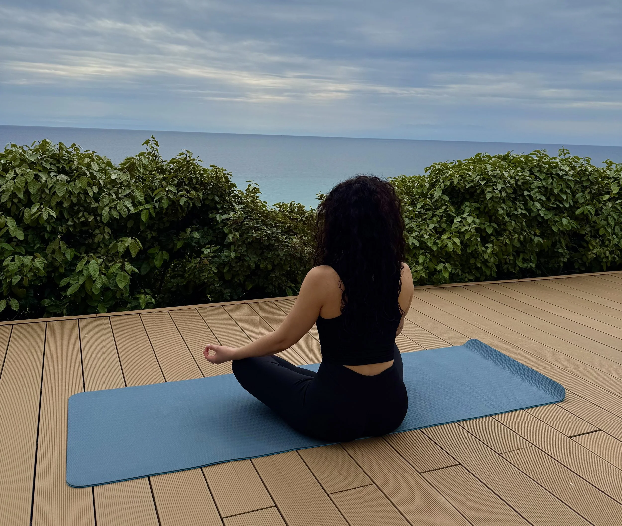 Woman sitting cross-legged on a yoga mat outdoors overlooking bushes and the ocean, practicing meditation or yoga.